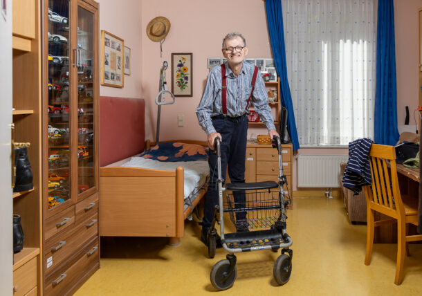 Ein Mann mit hellblauem Hemd, dunkelblauer Hose und roten Hosenträgern steht mit seiner Gehhilfe in seinem Zimmer und lächelt. Im Zimmer befindet sich ein Bett, ein Schreibtisch mit Sessel, ein Kästchen und eine Vitrine mit Miniatur-Autos.