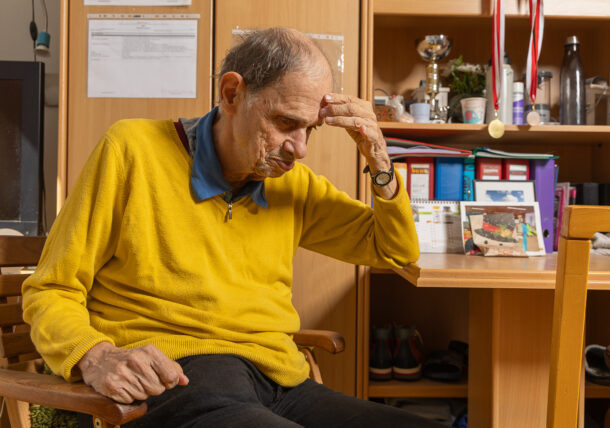 Ein Mann mit gelbem Pullover und blauen Kragen sitzt auf einem Sessel in seinem Zimmer. Das Regal hinter ihm ist voll mit persönlichen Dingen, Medaillen, Ordnern, Dosen, Schuhen.