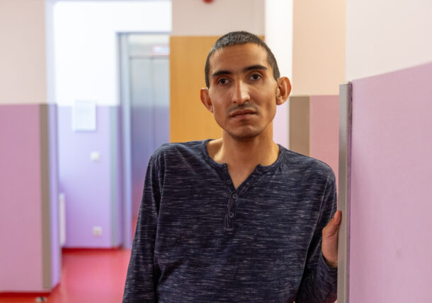 Ein junger Mann mit grauem Shirt und schwarzen Haaren steht in einem Gang mit lila gestrichenen Wänden und schaut in die Kamera.