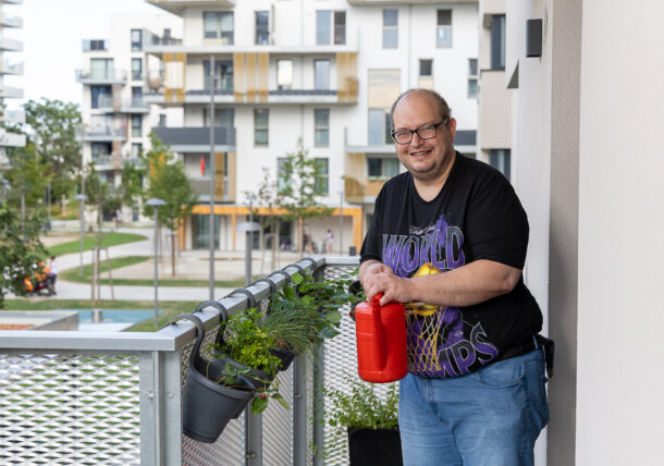 Auf dem Bild steht ein Mann mit kurzen Haaren, Brille und schwarzem T-Shirt mit einer roten Gießkanne auf dem Balkon seiner Wohnung. Am Balkon hängen Kräuter und im Hintergrund befinden sich Gebäude der Nachbarschaft.