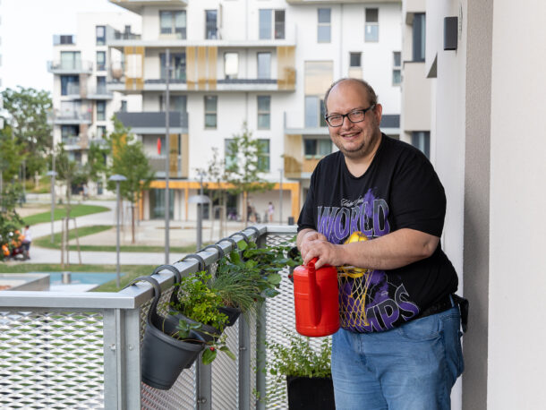Auf dem Bild steht ein Mann mit kurzen Haaren, Brille und schwarzem T-Shirt mit einer roten Gießkanne auf dem Balkon seiner Wohnung. Am Balkon hängen Kräuter und im Hintergrund befinden sich Gebäude der Nachbarschaft.
