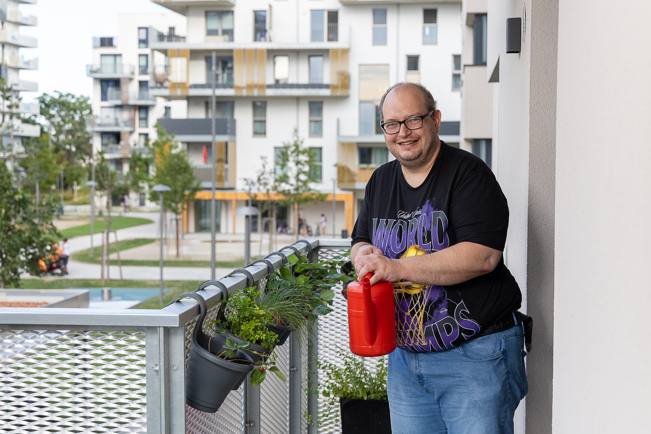 Auf dem Bild steht ein Mann mit kurzen Haaren, Brille und schwarzem T-Shirt mit einer roten Gießkanne auf dem Balkon seiner Wohnung. Am Balkon hängen Kräuter und im Hintergrund befinden sich Gebäude der Nachbarschaft.