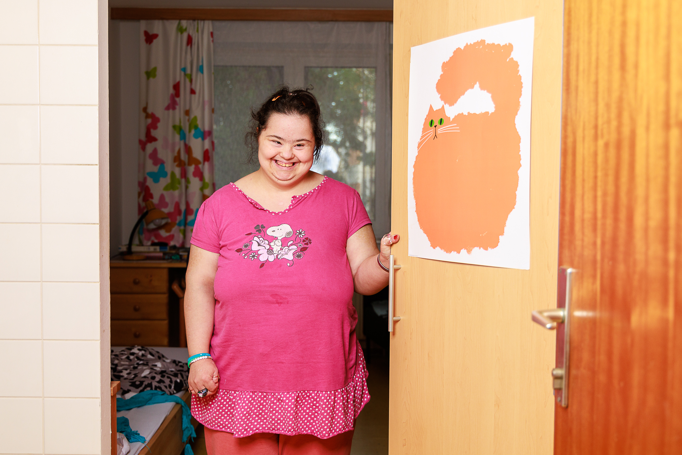 Eine lachende Frau mit hochgestecktem braunem Haar steht an der Tür zu ihrem Zimmer. Sie trägt ein pinkes Shirt mit Snoopy-Motiv. An der der Tür hängt ein Poster einer Katze als Comic.