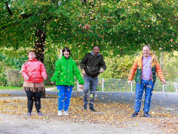 Vier Personen stehen lachend unter einem großen Baum und werfen Herbstlaub in die Luft. Im Hintergrund sind Bäume, ein Zaun und eine grüne Wiese zu sehen. Die Szene strahlt Freude und herbstliche Lebendigkeit aus.