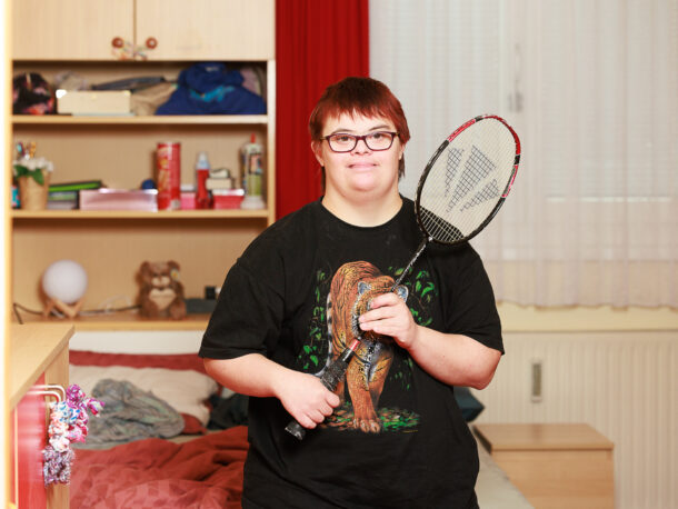 Eine Frau mit roten Haaren und Brille schaut freundlich in die Kamera. Sie trägt ein schwarzes T-Shirt mit Tiger-Motiv. Sie steht in ihrem Zimmer und hält einen Federball-Schläger in der Hand.