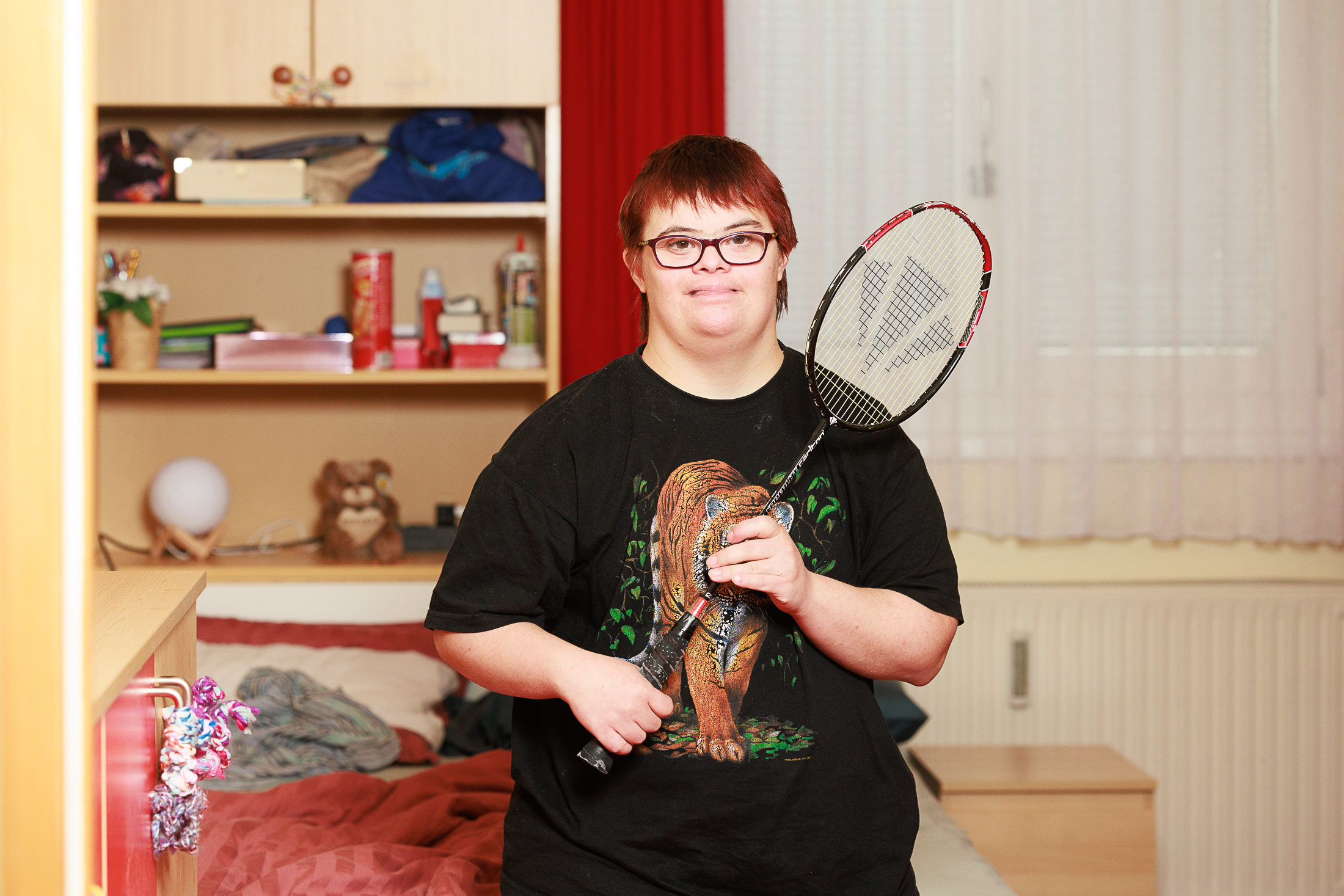 Eine Frau mit roten Haaren und Brille schaut freundlich in die Kamera. Sie trägt ein schwarzes T-Shirt mit Tiger-Motiv. Sie steht in ihrem Zimmer und hält einen Federball-Schläger in der Hand.