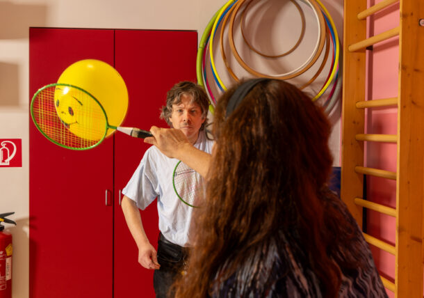 In einem Bewegungsraum mit Sprossenwand und Ringen in unterschiedlichen Farben und Größen an der Wand spielen ein Mann und eine Frau Badminton mit einem Luftballon, auf dem ein Smiley angebracht ist.