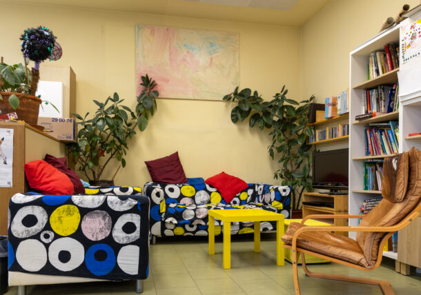 Das Foto zeigt einen Sofagarnituren mit bunten Stoffüberzug und einen Relaxsessel. An der Seite stehen Grünpflanzen, Buecherregale und in der Mitte ein gelber Couchtisch.