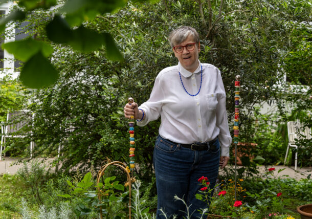 Das Foto zeigt eine Frau mit kurzen grauen Haaren, Brille, weißer Bluse und Jeans. Sie steht in einem gruenen Innenhof und stützt ihre Hand auf eine bunte Gartenstele mit bunten Keramikteilen. Im Hintergrund sieht man noch eine.