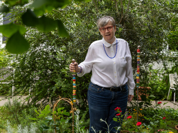 Das Foto zeigt eine Frau mit kurzen grauen Haaren, Brille, weißer Bluse und Jeans. Sie steht in einem gruenen Innenhof und stützt ihre Hand auf eine bunte Gartenstele mit bunten Keramikteilen. Im Hintergrund sieht man noch eine.