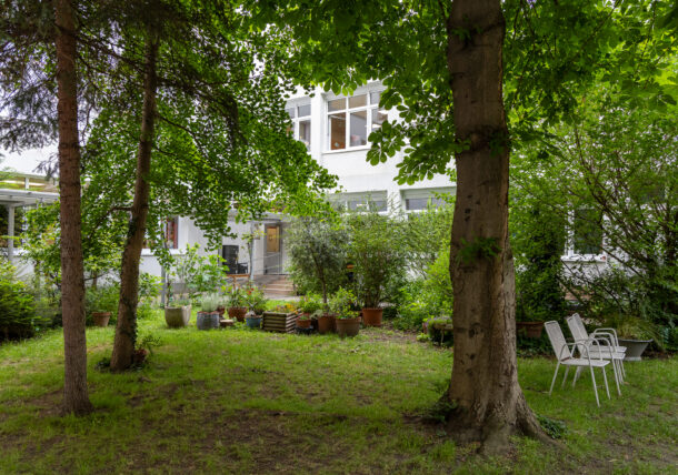Das Foto zeigt einen grünen Garten mit Bäumen, Strauechern, Toepfen. Man sieht weiße Gartensessel und im Hintergrund die weiße Fassade der Rauemlichkeiten der Tagesstruktur