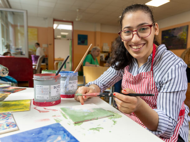 Eine junge Frau mit Brille und gestreifter Bluse und Schürze sitzt in der Kreativgruppe der Tagesstruktur. Sie hat einen Pinsel in der Hand, Farben vor sich stehen und bemalt eine Fliese. Sie hat lange schwarze Haare und laechelt freundlich in die Kamera.