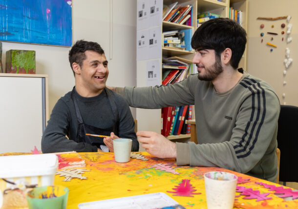 Das Foto zeigt zwei juengere Maenner, sie lachen sich freundlich an, einer legt seine Hand auf die Schultern des anderen und sie bemalen Christbaueme aus Holz. Sie sitzen an einem Tisch in der Tagesstruktur.