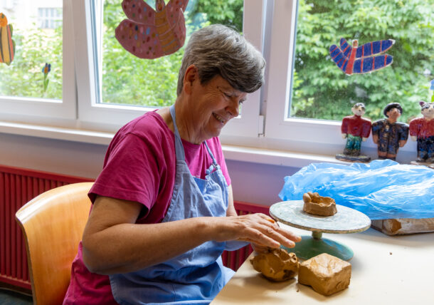 Das Foto zeigt eine Frau mit grauen, kurzen Haaren, Sie trägt ein pinkes T-Shirt und eine blaue Arbeitsschuerze. Sie sitzt an einem Tisch und fertigt eine Skulputur aus Ton an. Vor ihr liegt ein Block aus Ton. Sie hat einen Fensterplatz in der