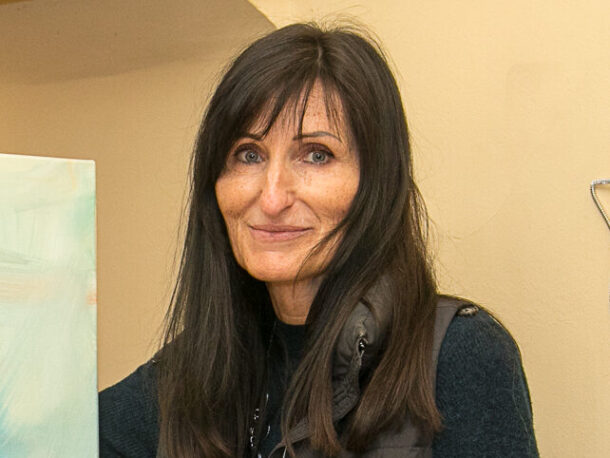 Die Mitarbeiterin Monika Trummer schaut in die Kamera. Sie steht neben einem Bild auf einer Staffelei, hat lange schwarze Haare und trägt ein dunkles Strickkleid mit Gilet.