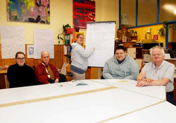 Eine Personengruppe sitzt an einem Tisch zusammen. Sie lesen und schauen sich Zeitungen an. Hinter ihnen schreibt eine Frau auf einem Flipchart, daneeben steht ein Mann, ein weiteres Mitglied der Zeitungsgruppe.
