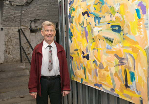 Ein älterer Mann mit grauem Haar, mit weinroter Strickjacke, weißem Hemd und Krawatte laechelt in die Kamera. Neben ihm haengt ein selbtgemaltes großes Bild an der Wand.