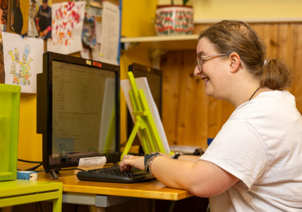 Das Foto zeigt eine Frau mit Brille, zusammengebundenen blonden Haaren, weißem T-Shirt. Sie sitzt an einem Schreibtisch in der Tagesstruktur und tippt auf einer Tastatur und sieht in den Bildschirm.