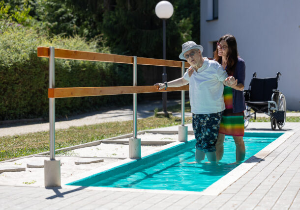 Eine Frau mit Hut, weißem T-Shirt und blauer Hose und eine Frau mit buntem Sommerkleid und Weste stehen in einem flachen Wasserbecken. Neben ihnen ist ein Geländer, an dem man sich festhalten kann. Im Hintergrund ist ein Rollstuhl zu sehen.