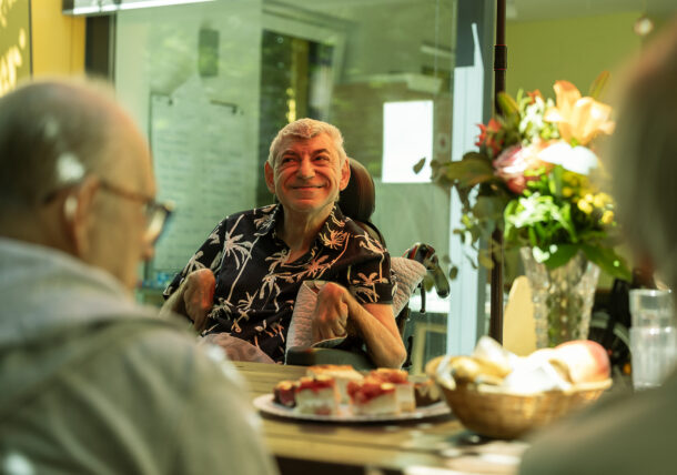 Ein Mann mit schwarzem Hemd mit Palmenmusterr sitzet am Tisch. Er ist Rollstuhlfahrer. Am Tisch stehen eine Vase mit einem Blumsenstrauß und Gebäck. Im Hintergrund sieht man den Aufenthaltsraum.