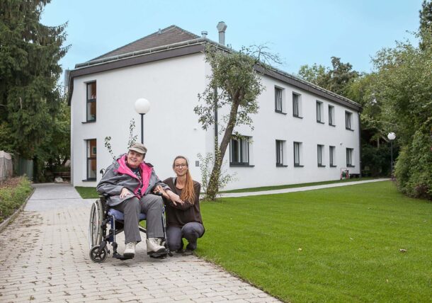 Zwei Frauen posieren vor einem Wohnhaus mit Garten. Die Frau links sitzt im Rollstuhl, die Mitarbeiterin kniet rechts neben ihr.