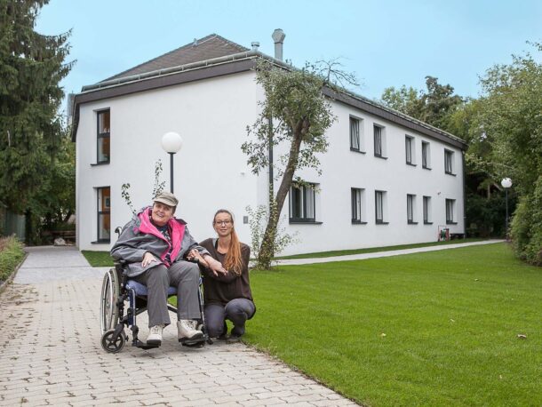 Zwei Frauen posieren vor einem Wohnhaus mit Garten. Die Frau links sitzt im Rollstuhl, die Mitarbeiterin kniet rechts neben ihr.