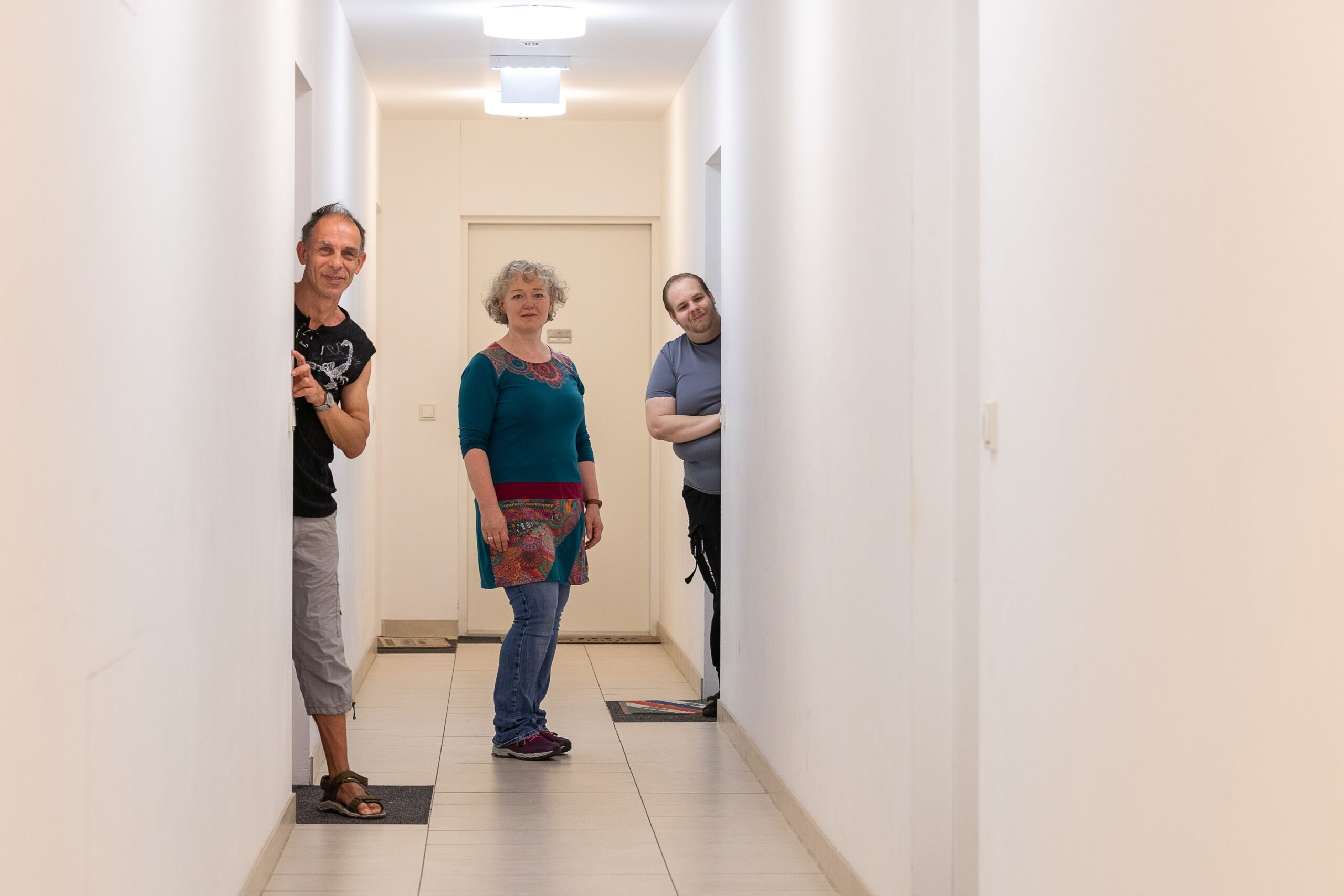 Zwei Männer schauen aus ihren Wohnungstüren. Am Gang in der Mitte steht die Assistentin der Lebenshilfe Wien. Sie ist eine Frau mit grauem mittellangem Harren und trägt ein türkises Kleidl. Sie unterstützt die beiden Kunden der Lebenshilfe Wien im Alltag in ihren eigenen Wohnungen.