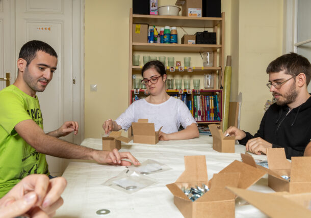Mehrere Personen sitzen an einem Tisch und verpacken Artikel einer Firma in eine Kartonbox. Die Gruppe arbeitet konzentriert.