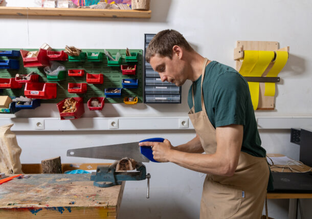 Man sieht einen jungen Mann mit brauner Arbeitschuerze von der Seite. Er ist damit beschaeftigt aus einem Holzteil eine Scheibe abzusaegen. Er arbeitet in der Holzwerkstatt in der Tagesstruktur in der Schottengasse.