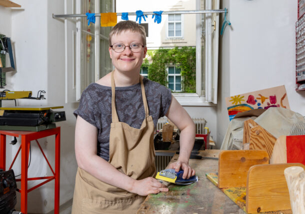Eine junge Frau mit Brille und brauner Arbeitsschuerze arbeitet in der Holzgruppe, sie schleift gerade.