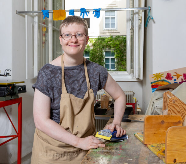 Eine junge Frau mit Brille und brauner Arbeitsschuerze arbeitet in der Holzgruppe, sie schleift gerade.