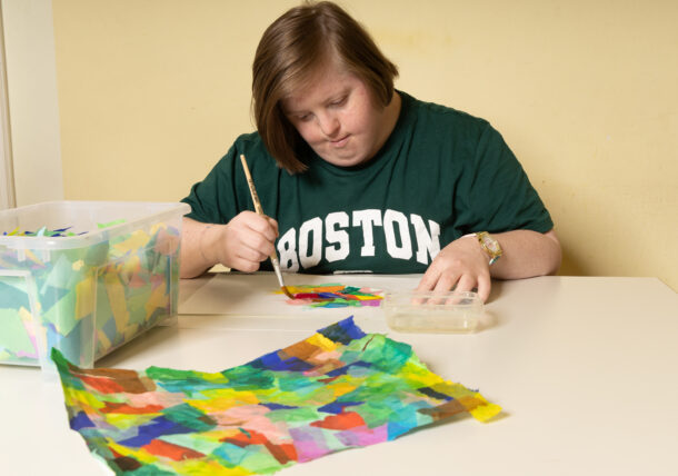 Eine Frau mit Down Syndrom sitzt an einem Tisch und leimt mit einem Pinsel Papierstücke aneinander. Vor ihr liegt ein fertiges bunten Arbeitsstück. Sie traegt ein grünes T-Shirt mit der Aufschriftt BOSTON, neben ihr steht eine Kiste mit Papierschnipsel und vor ihr eine kleine Dose mit Leim.