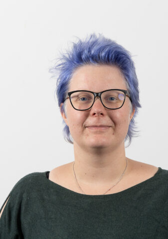 Die Mitarbeiterin Julia Scamoni schaut freundlich in die Kamera. Sie leitet das Garconnierenwohnen der Lebenshilfe Wien. Sie hat blaue Haare, trägt eine Brille und grünes Oberteil.