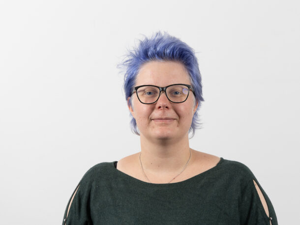 Die Mitarbeiterin Julia Scamoni schaut freundlich in die Kamera. Sie leitet das Garconnierenwohnen der Lebenshilfe Wien. Sie hat blaue Haare, trägt eine Brille und grünes Oberteil.