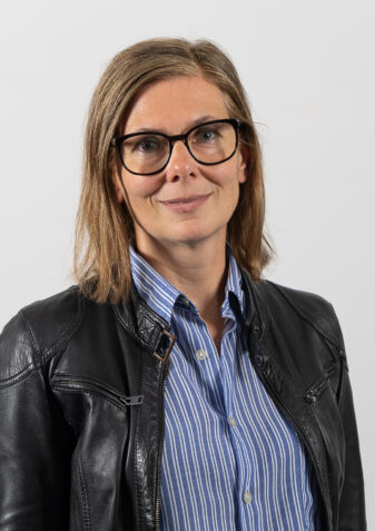 Die Fachliche Leitung Karin Gerbautz laechelt in die Kamera. Sie hat schulterlange braune Haare und traegt eine Brille. Sie hat eine schwarze Lederjacke an und darunter eine blau gestreifte Bluse.