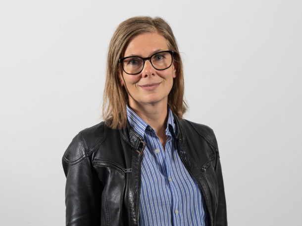 Die Fachliche Leitung Karin Gerbautz laechelt in die Kamera. Sie hat schulterlange braune Haare und traegt eine Brille. Sie hat eine schwarze Lederjacke an und darunter eine blau gestreifte Bluse.