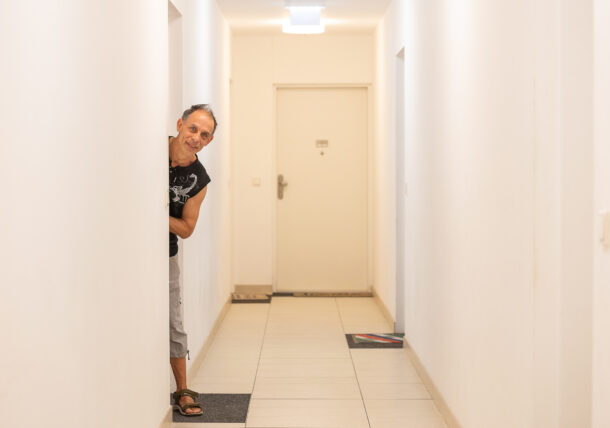 Ein Mann schaut freundlich aus seiner Wohnungstuere heraus. Einen Fuß hat er auf seiner Tuermattte stehen. Im Hintergrund sieht man weitere Wohnungstüren in einem weiß ausgemalten Gang einer Wohnhausanlage.
