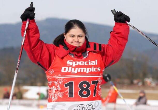 Eine Langlauferin mit Down Syndrom streckt ihre Haende mit ihren Stoecken in die Luft. Sie hat lange zusammengebundene dunkle Haare und trägt einen roten Anorak und ein Nummern-Shirt mit der Aufschrift Special Olympics und der Zahl 187.