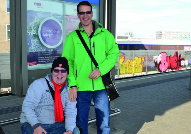 Zwei Männer sitzen auf einem Bahnsteig und lachen in die Kamera.