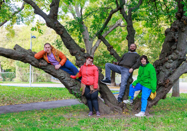 Eine Gruppe von zwei Frauen und Männer stehen vor und in einem großen Baum in einer Parkanlage. Die beiden Männer lehnen an den großen Baumstämmen und die Damen stehen vor ihnen. Alle schauen freundlich in die Kamera und haben Outdoorkleidung an.