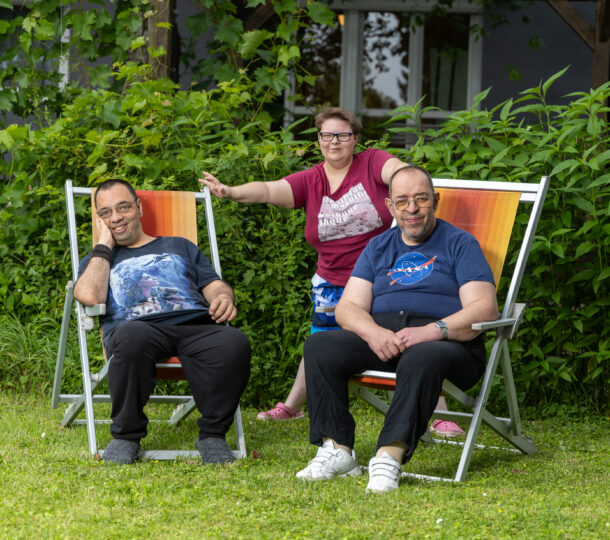 Das Foto zeigt zwei Maenner und eine Frau in einem Garten vor einem Wohnhaus der Lebenshilfe. Die beiden Herren sitzen in einem Liegestuhl, die Dame steht in der Mitte hinter ihnen.
