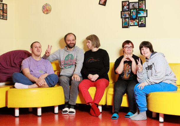 Eine Gruppe von fuenf Menschen mit Behinderungen sitzt froehlich zusammen auf einer gelben Couch in einem Wohnzimmer. Hinter ihnen an der Wand haengen Fotos.