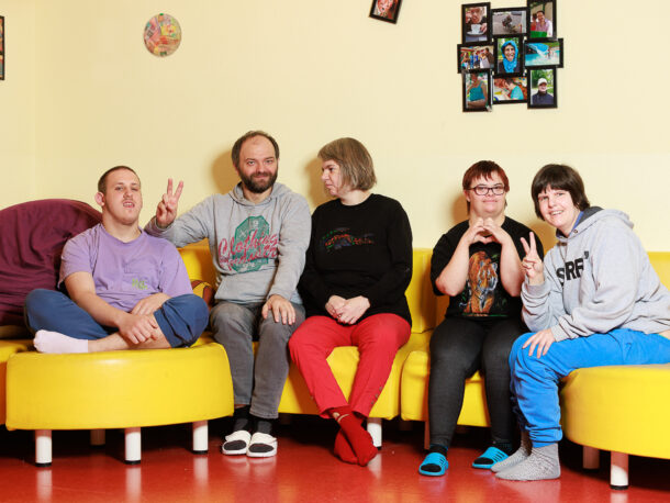Eine Gruppe von fuenf Menschen mit Behinderungen sitzt froehlich zusammen auf einer gelben Couch in einem Wohnzimmer. Hinter ihnen an der Wand haengen Fotos.