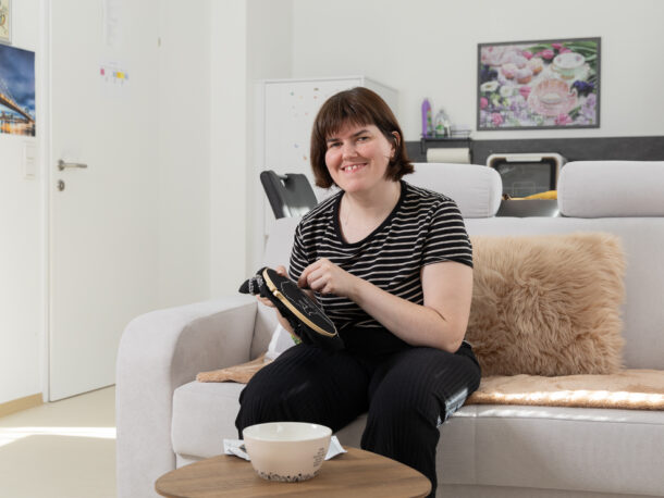 Das Bild zeigt eine Frau mit kurzen braunen Haaren beim Sticken. Sie sitzt auf einer Couch und schaut zufrieden in die Kamera. Das Zimmer ist freundlich und hell eingerichtet.