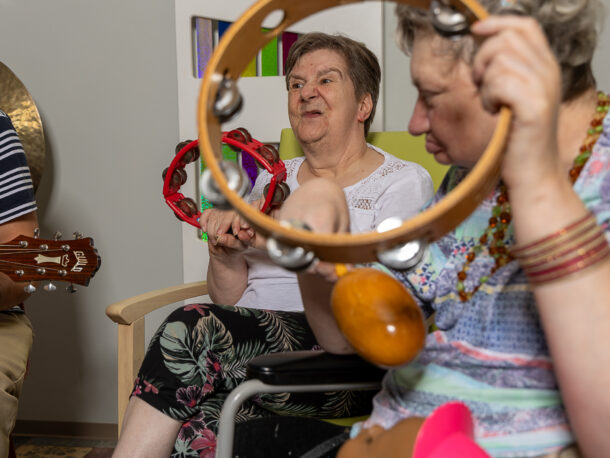Das Bild zeigt zwei Frauen mit Tamburin, die gemeinsam musizieren. Beide sitzen in einem Musikzimmer, umgeben von Musikinstrumenten. Die Atmosphäre ist entspannt und kreativ.