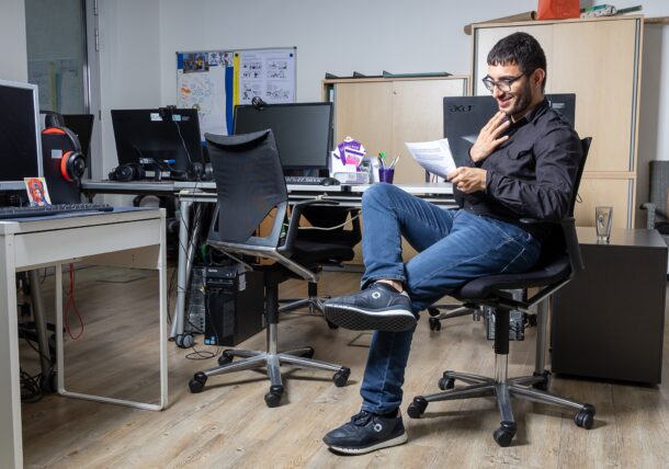 Ein junger Herr mit Brille sitzt auf einem Bürosessel in einem Büro. Er lächelt und liest einen Text.