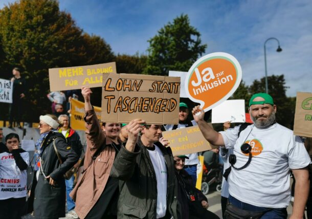 Eine Gruppe KundInnen der Lebenshilfe demonstrieren. Sie halten auf einem Platz in der Wiener Innenstadt Schilder mit ihren Forderungen hoch. Darauf steht zum Beispiel: Ja zur Inklusion und Lohn statt Taschengeld.