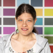 Die Kundin Natalie Hajek schaut freundlich in die Kamera. Sie hat dunkle zusammengebundene Haare, steht vor einer bunten Wand und hat ein weiß gemustertes Oberteil an.