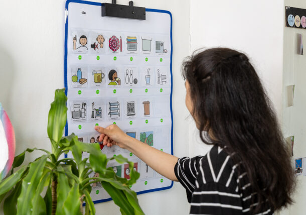 Eine junge Dame drückt auf ein Symbol auf einer Wandtafel. Es handelt sich dabei um eine sogenannte Sprechende Wand, die ein Hilfsmittel der Unterstützten Kommunikation ist.
