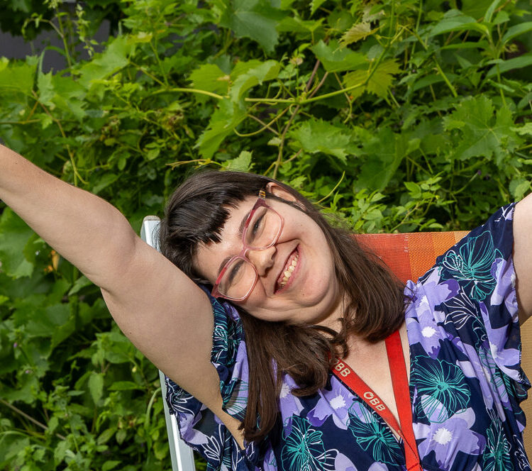 Junge Frau mit langen dunklen Haaren sitzt in einem Liegestuhl im Garten. Sie lächelt fröhlich in die Kamera und steckt beide Hände in die Höhe.